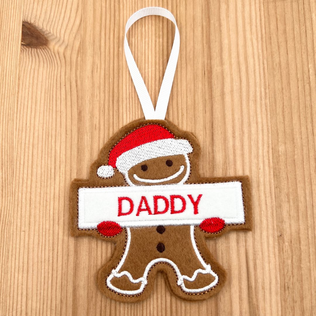 A gingerbread Dad felt ornament hanging on a light wood background. The ornament is brown with a white hanging loop, a red and white Santa hat, red mittens, and a white banner across the chest that says “DADDY” in red letters.
