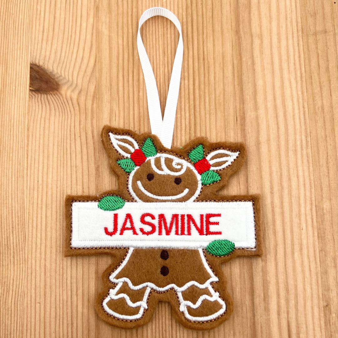 A personalized gingerbread girl ornament hanging on a light wood background. The ornament features a brown gingerbread figure with white icing details, green and red holly accents, and a white banner across the chest with the name 'JASMINE' in red embroidered letters.