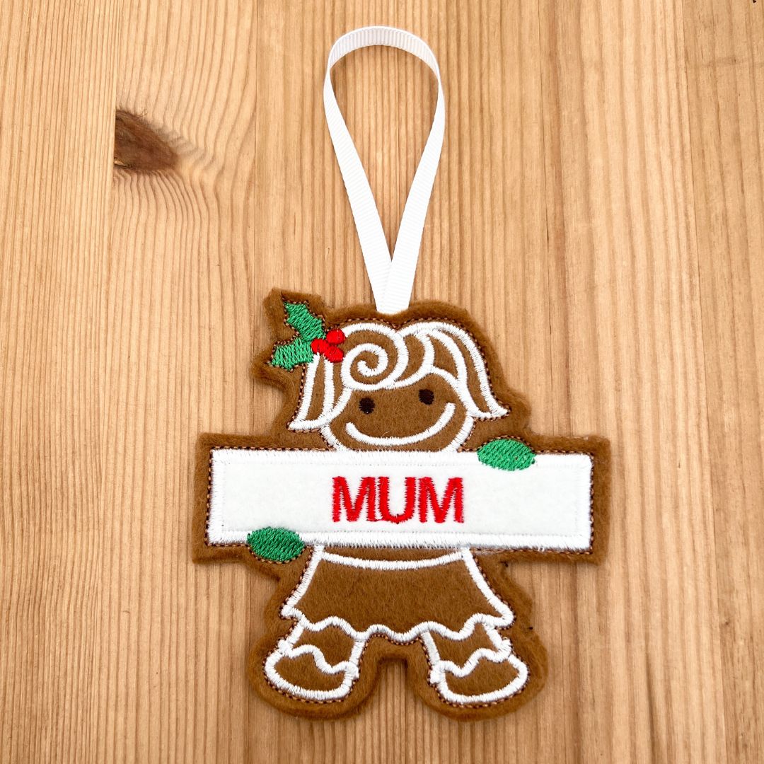 A personalized felt Christmas Gingerbread Mum ornament shaped like a gingerbread person holding a white banner that says 'MUM' in red letters. The ornament has a white hanging ribbon and is displayed on a light wood background.