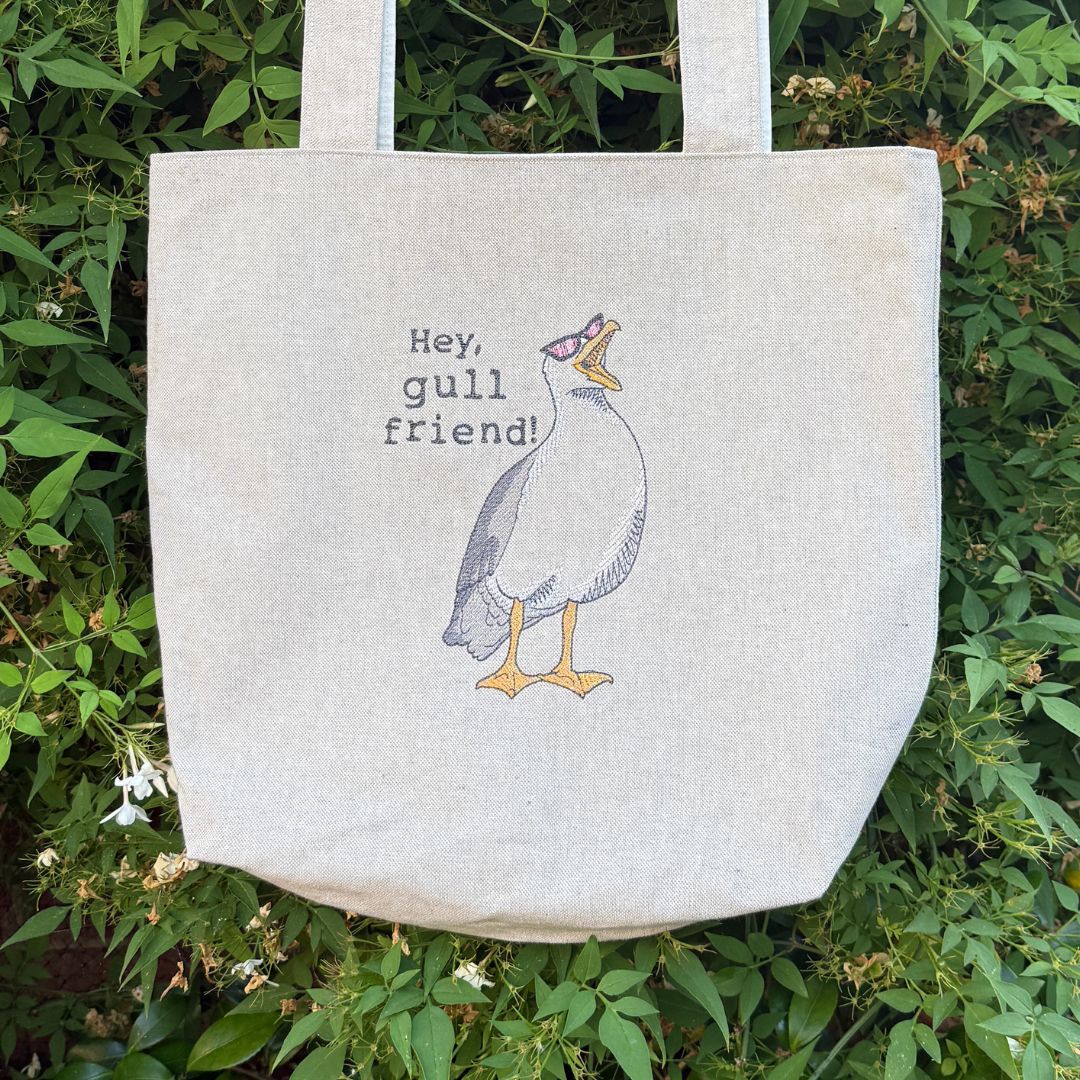 A natural linen tote bag with an embroidered illustration of a gull wearing sunglasses and the text 'Hey, gull friend!' displayed outdoors against a background of green foliage.