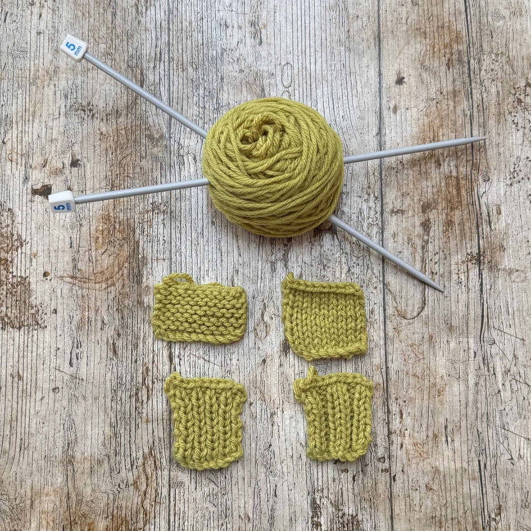 A ball of olive green yarn on gray knitting needles, arranged on a rustic wooden surface with four small knitted swatches below to show stitch texture and color.