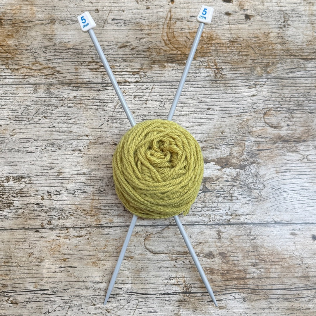 A ball of olive green yarn wrapped around two gray knitting needles with white tips marked '5', placed on a rustic weathered wood background.