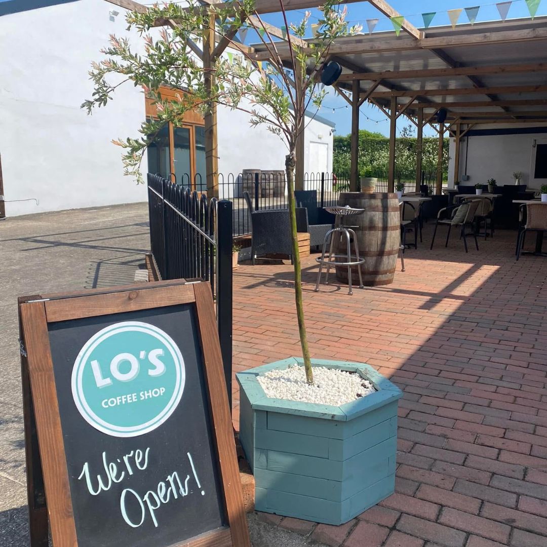 Outdoor seating area with a 'LO'S Coffee Shop' sign indicating 'We're Open!' on a sunny day.