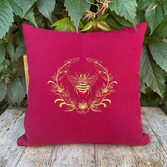 Red pillow with gold bee and floral design on a wooden surface with green leaves in the background