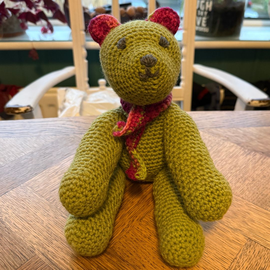 A handmade olive green crocheted teddy bear with pink ears and a red patterned crocheted scarf, sitting on a wooden table in a home setting. completed in a Level 2 beginners Crochet workshop with Coralie's Crafts & Gifts