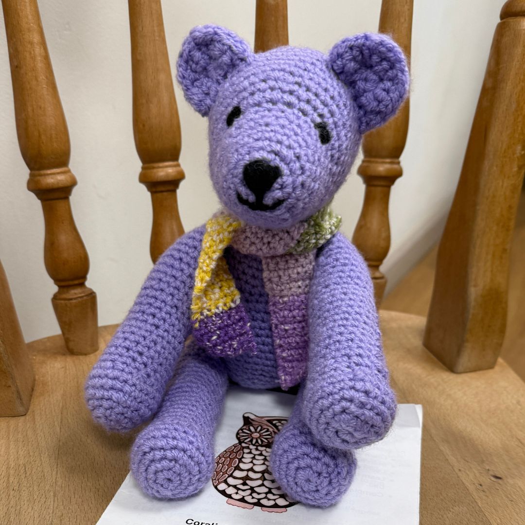 A handmade purple crocheted teddy bear wearing a yellow, purple, and green striped scarf, sitting on a wooden chair with the Level 2 beginners crochet workshop booklet underneath, completed at Coralie's Crafts & Gifts.