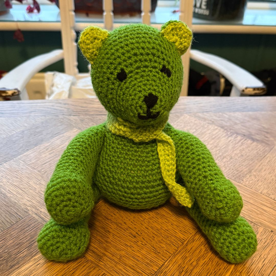 A handmade green crocheted teddy bear with a yellow-green scarf sits on a wooden table in a home setting, made during private Level 2 Beginners Crochet  workshop, with Coralie's Crafts & Gifts.