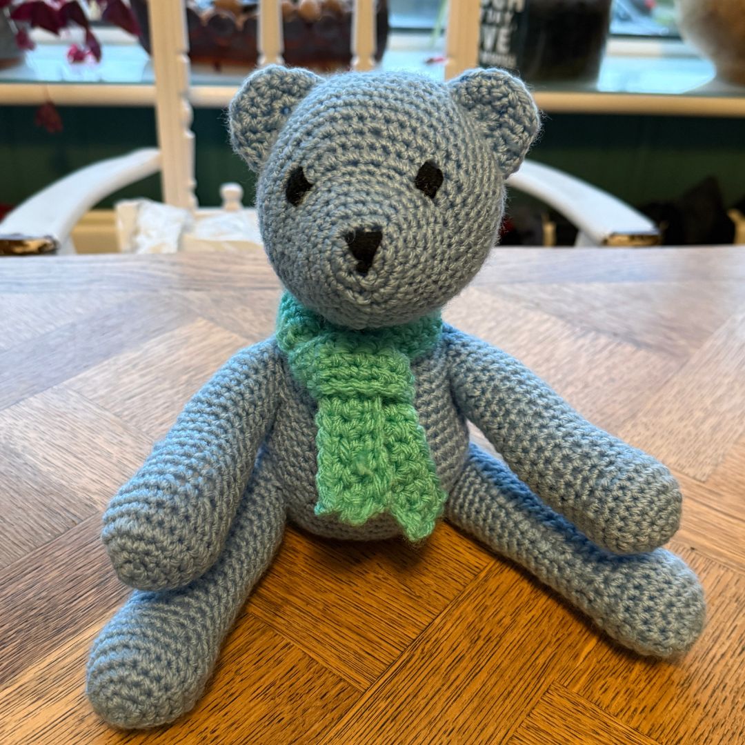 A crocheted teddy bear sitting on a wooden table, made from light blue-gray yarn with a green knitted scarf and small black embroidered eyes and nose, made whilst attending a Private workshop completing Level 2 Beginners Crochet Workshop in Dorset with Coralie's Crafts & Gifts.