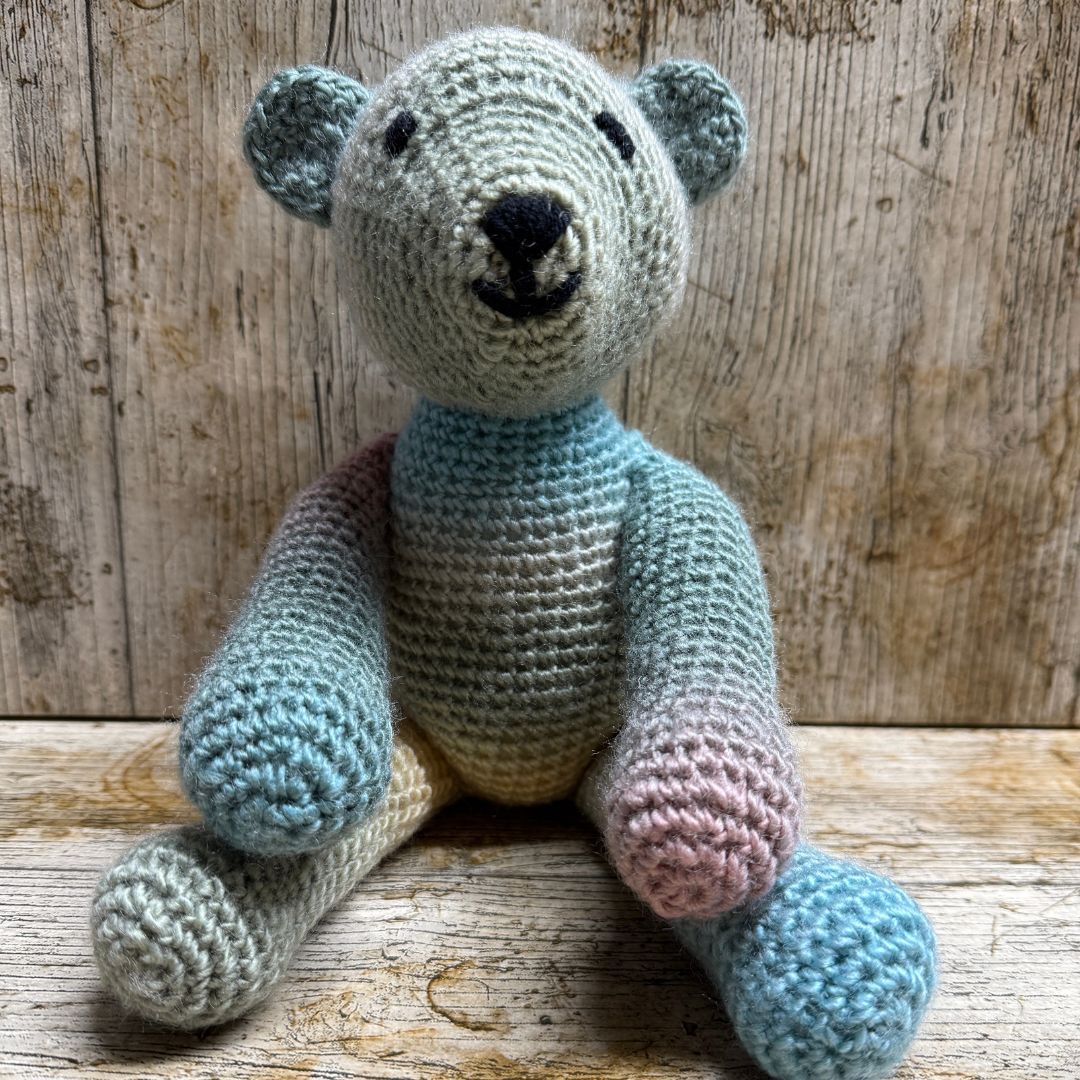 A handmade crocheted teddy bear toy with a color-changing gradient design, sitting on a rustic wooden surface against a weathered wood plank background, made whilst attending a Level 2 Beginners Crochet workshop, with Coralie's Crafts & Gifts