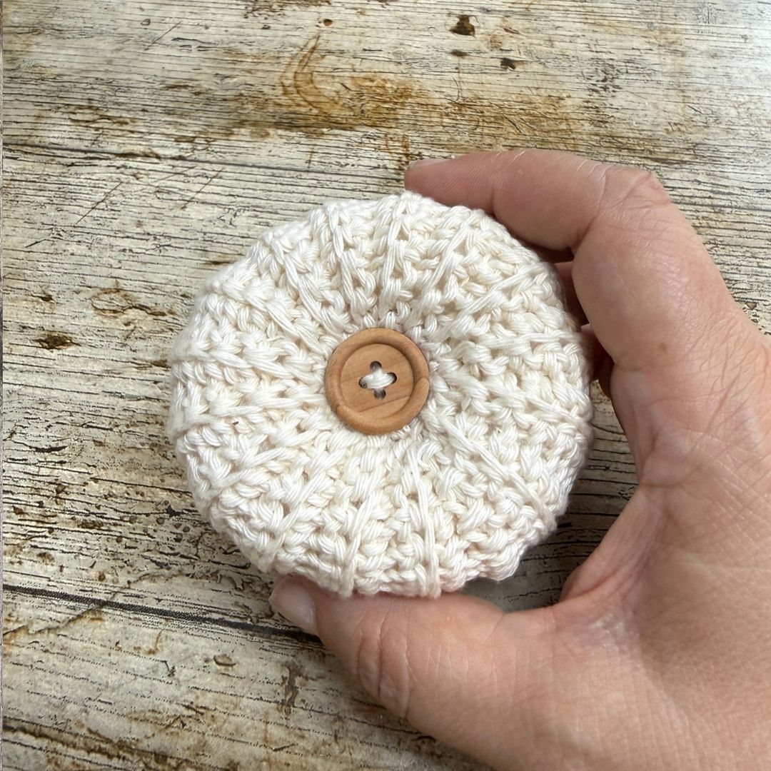 A hand holding a small round, off-white crocheted fabric item with a tan button in the center, set against a rustic wooden surface.