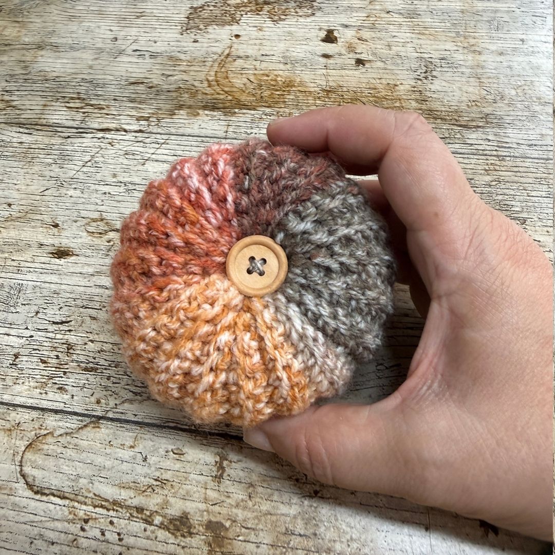 A hand holding a small round crocheted item with a button on top, featuring a variegated color pattern of orange, pink, brown, and gray, set against a rustic wooden background.