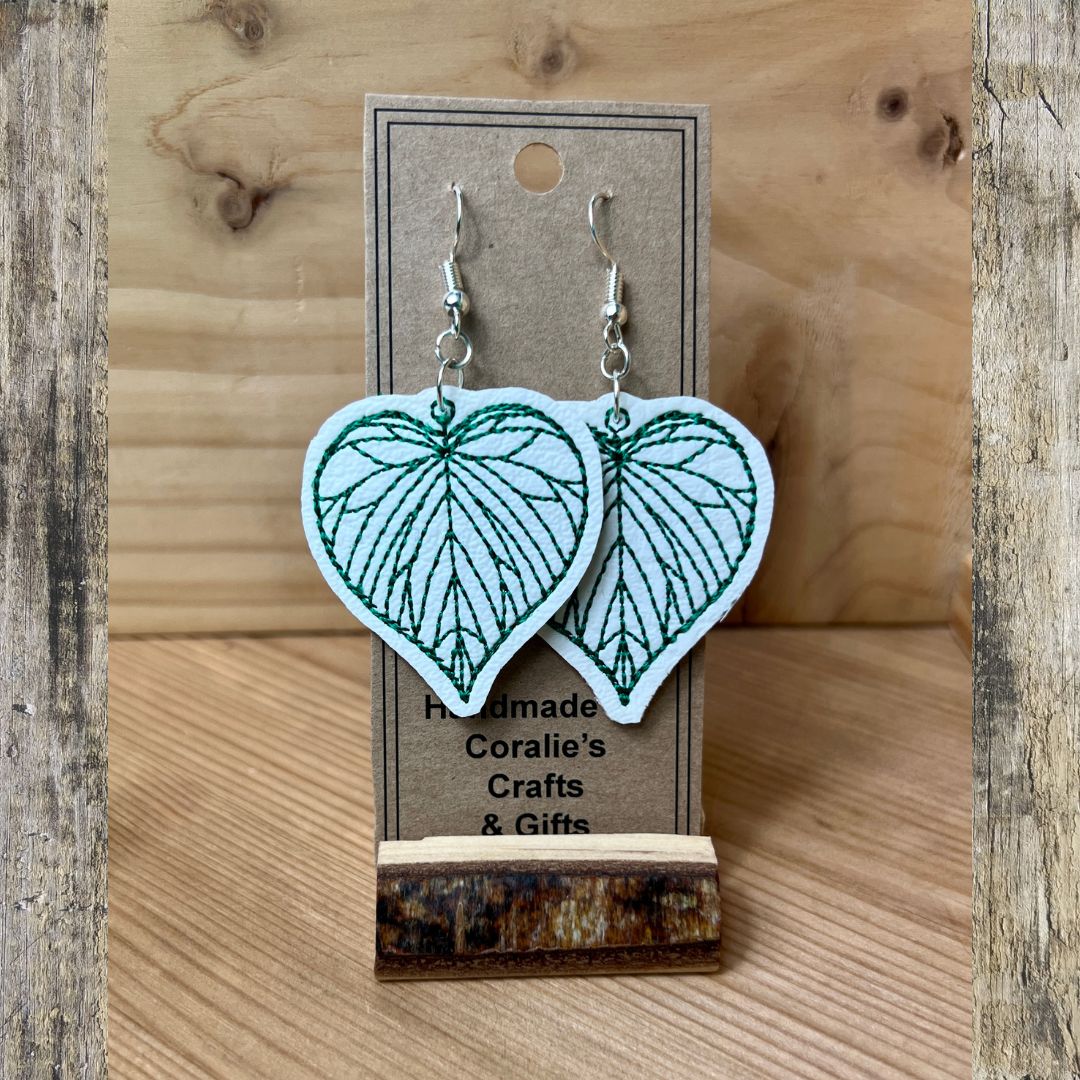 A pair of heart-shaped dangle earrings with a white fabric background and green leaf vein embroidery, hanging from silver hooks on a brown kraft display card. The card is mounted on a rustic wooden block over a light wood tabletop.