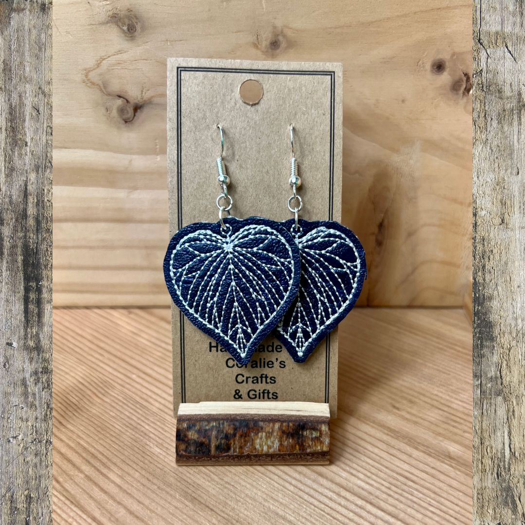 A pair of heart-shaped embroidered dangle earrings in navy blue and white, hanging from silver fishhook wires on a brown kraft display card. The card is set against a rustic wooden background.