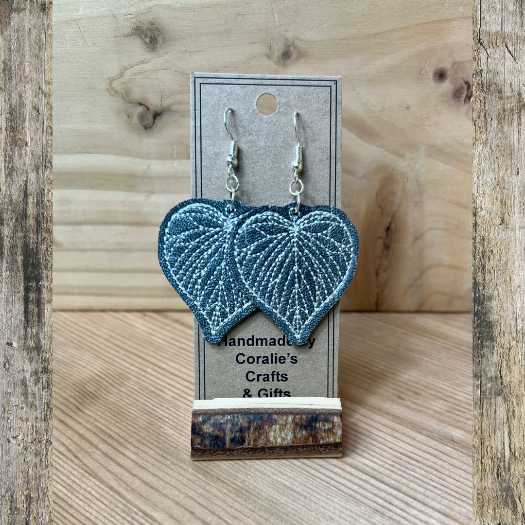A pair of heart-shaped dangle earrings with embroidered blue leaves, hanging from silver fishhook wires on a gray display card. The card is mounted on a small wooden block and set against a light wood grain background framed by rustic wood boards.