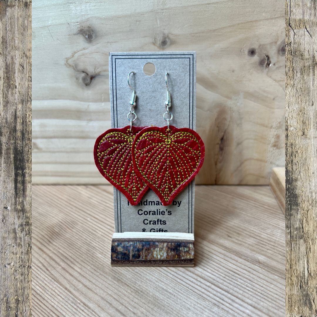 A pair of red heart-shaped dangling earrings with gold beaded detailing, displayed on a gray card labeled 'Handmade by Coralie's Crafts & Gifts' on a wooden surface.