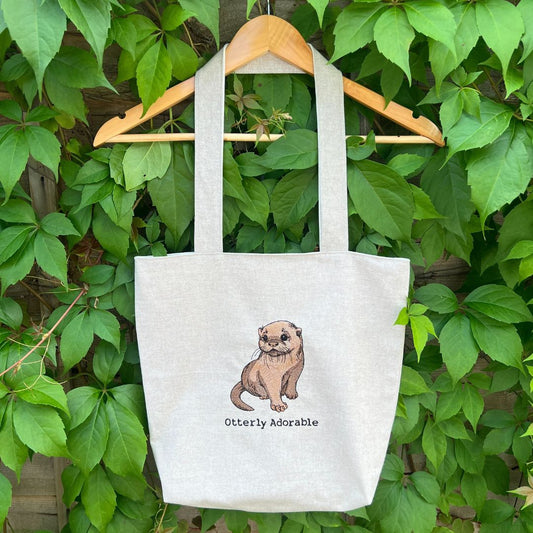 Tote bag with an otter illustration and 'Otterly Adorable' text, hanging on a wooden hanger against a green leafy background.