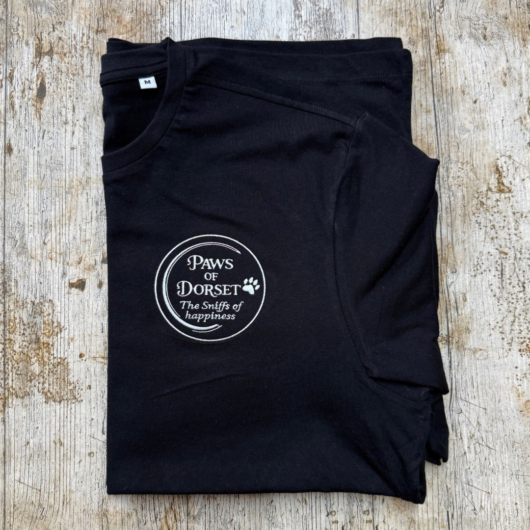 A folded black t-shirt with a white circular graphic reading 'Paws of Dorset The smiles of happiness' and a paw print, laid flat on a rustic light wood background.