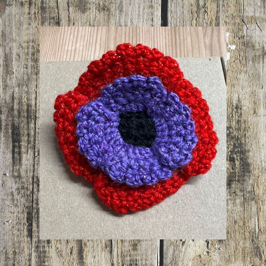 Crocheted flower poppy with red border and purple in the middle on a wooden surface