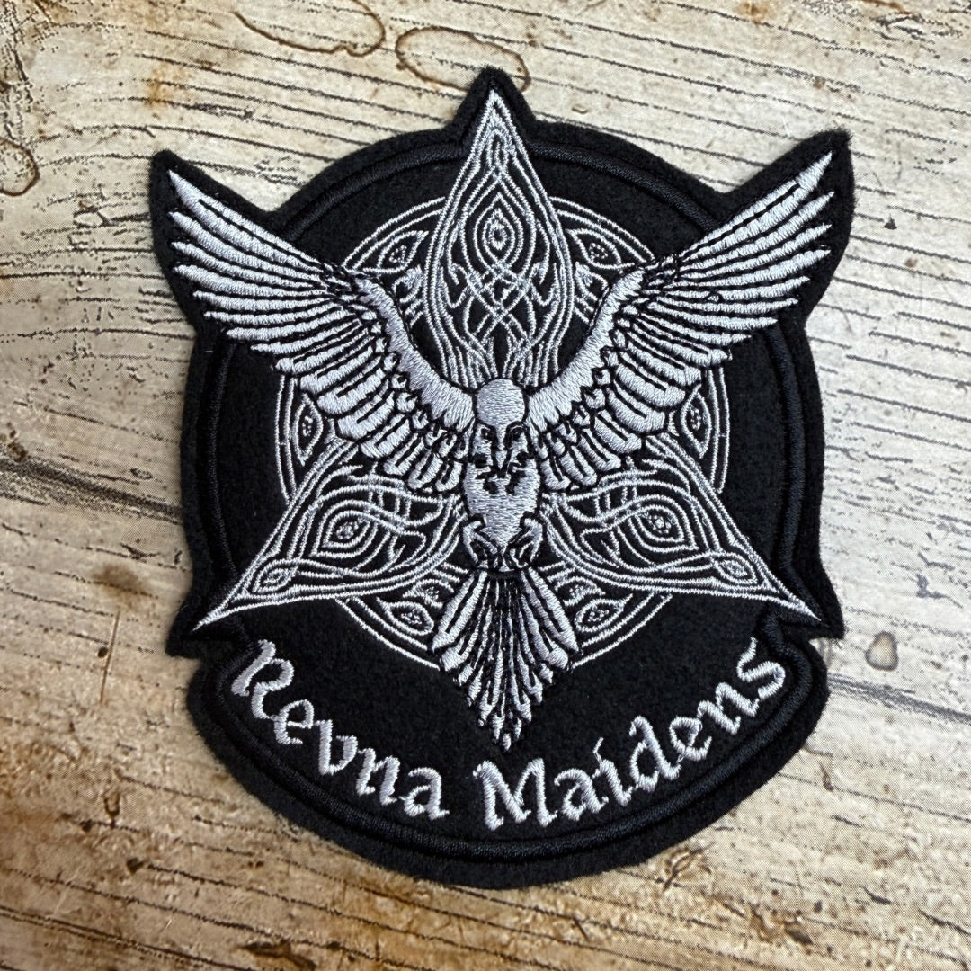 A circular embroidered patch featuring a black background with a silver-white winged emblem and the text 'Revna Maidens' along the bottom, placed on a rustic wooden surface.