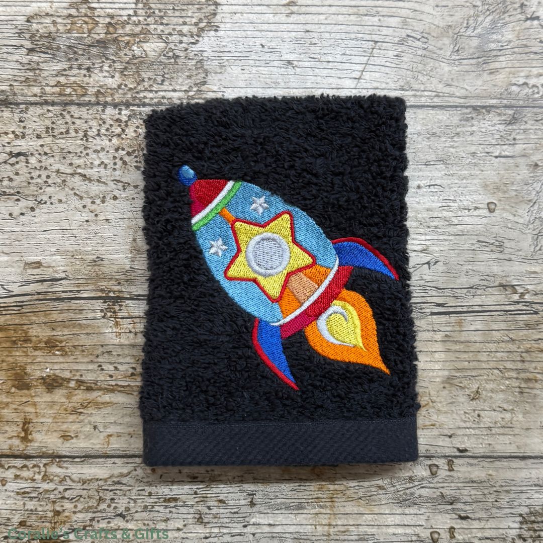 Black embroidered towel with a colorful rocket design on a wooden surface