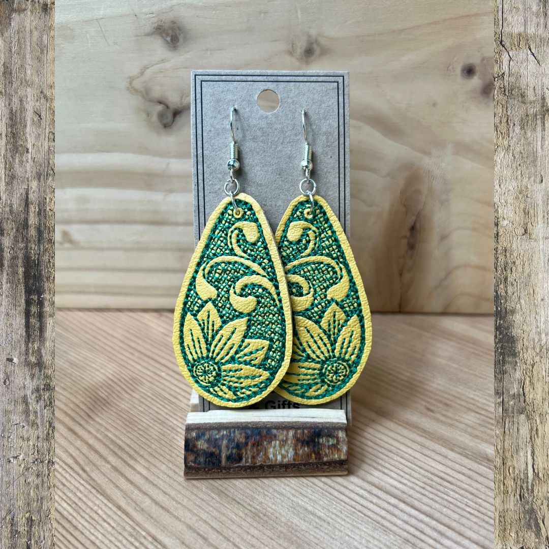 A pair of yellow embroidered teardrop-shaped dangle earrings, with  a green embroidered flower displayed on a gray card stand against a rustic wood background.