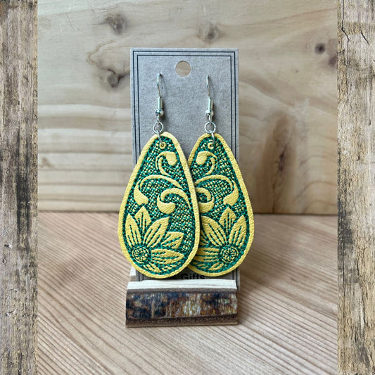 A pair of yellow embroidered teardrop-shaped dangle earrings, with  a green embroidered flower displayed on a gray card stand against a rustic wood background.