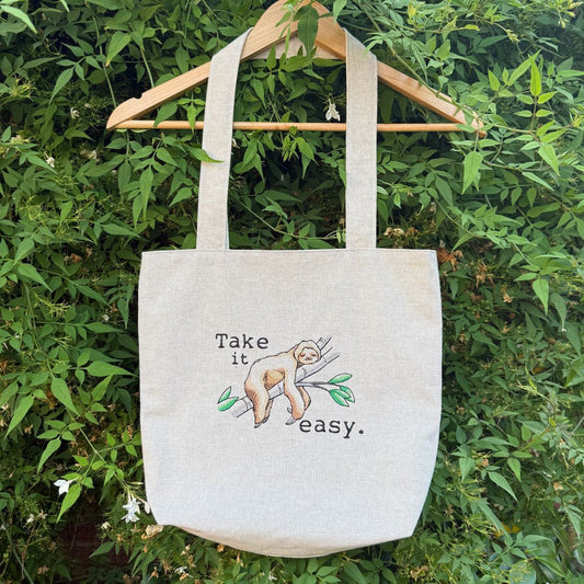 A natural canvas tote bag hanging on a wooden hanger against a background of green foliage. The bag features an embroidered sloth on a branch with the text 'Take it easy.