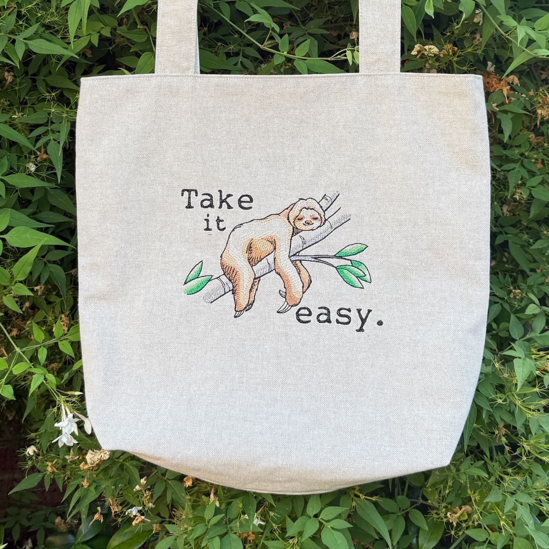 A natural canvas tote bag with an embroidered sloth illustration and the text 'Take it easy.' is photographed outdoors against a background of green foliage.
