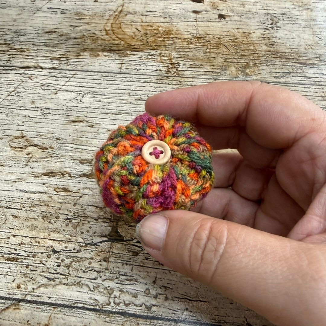 A small hand-crocheted multicolor pom-pom with a decorative button on top, held in a person's hand over a rustic wooden surface.