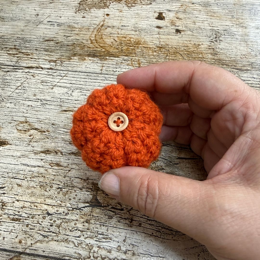 A small orange crocheted pumpkin-shaped accessory with a light beige button on top, held in a person's hand over a rustic whitewashed wooden surface.