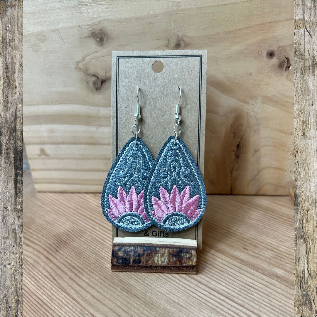 A pair of embroidered teardrop-shaped dangle earrings displayed on a small card stand. The earrings are light blue with pink lotus flower embroidery and silver fishhook wires.