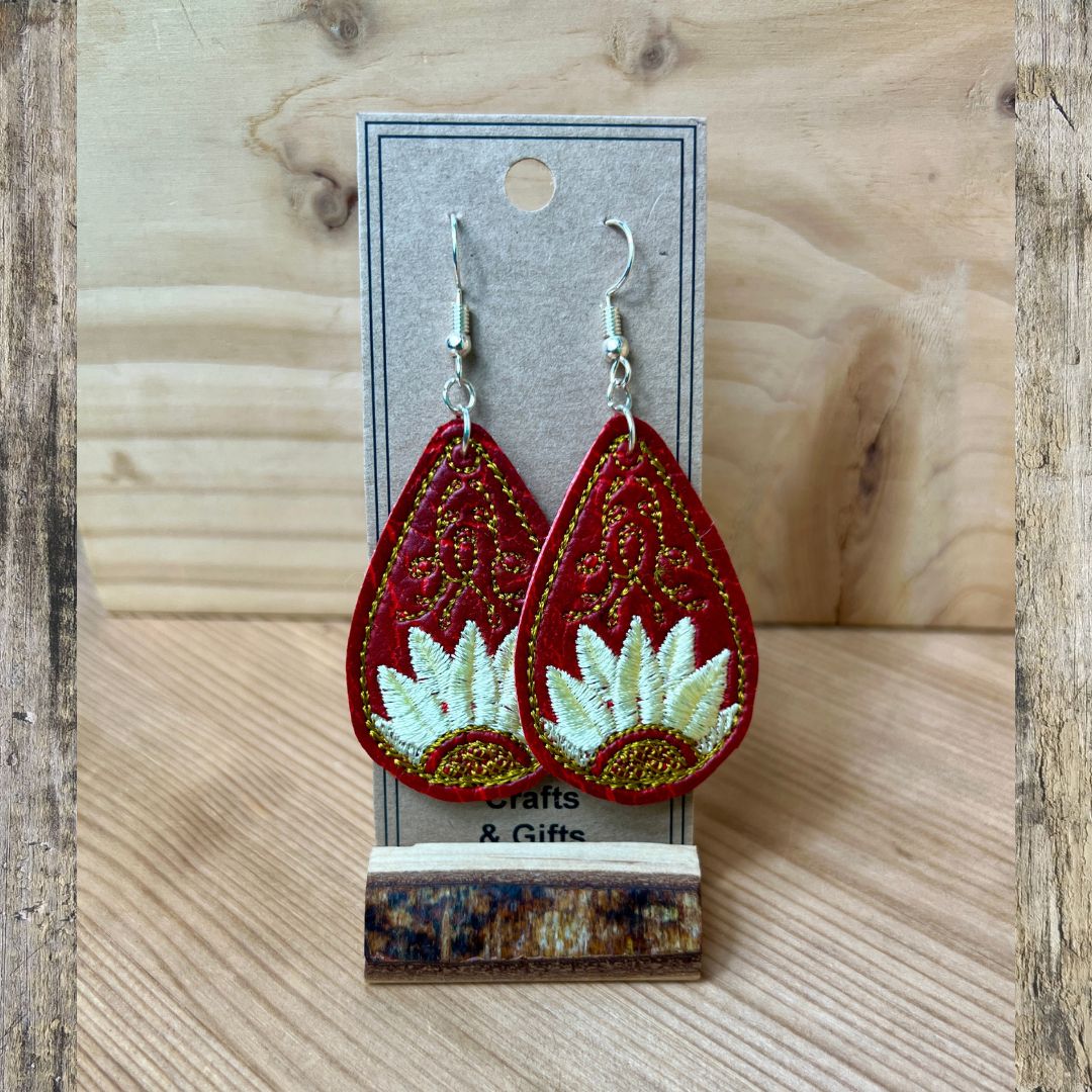 A pair of red teardrop-shaped embroidered earrings with white floral embroidery and gold glitter accents, displayed on a gray card labeled 'Crafts & Gifts' against a light wood background.