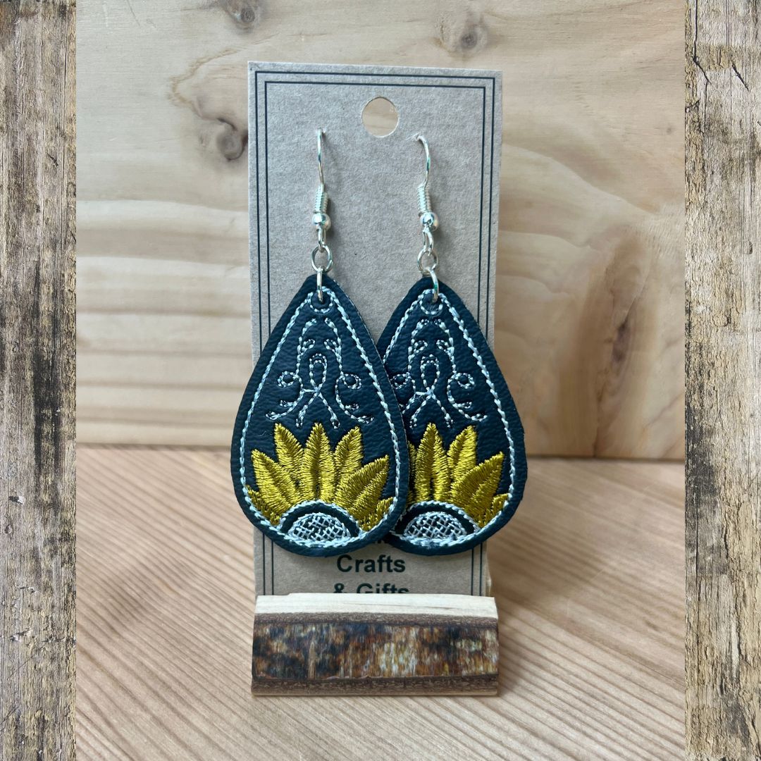A pair of teardrop-shaped embroidered earrings displayed on a gray card stand. The earrings are dark teal with white embroidered patterns and a yellow sunburst design in the center, hanging from silver fishhook wires.