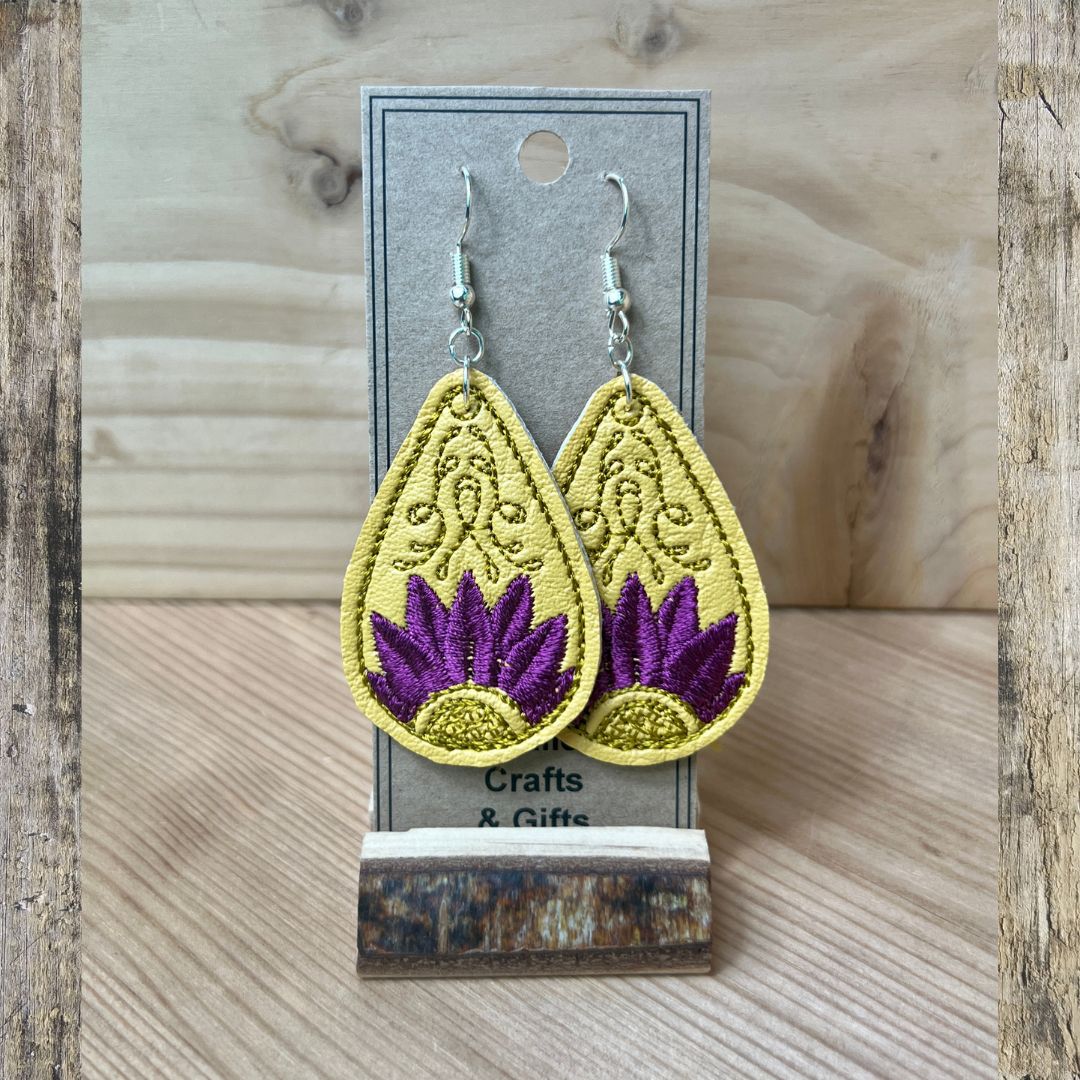 A pair of teardrop-shaped dangle earrings displayed on a gray card stand. The earrings have a yellow embroidered background with a purple lotus flower design and silver fishhook wires.