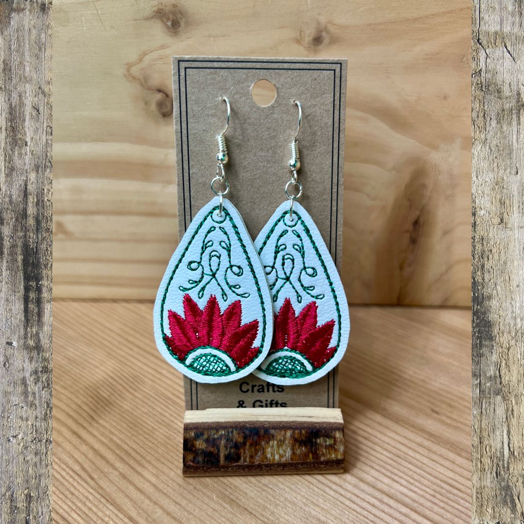 A pair of embroidered teardrop-shaped dangle earrings displayed on a brown card stand. The earrings have a white background with green embroidered scrollwork and a red embroidered lotus flower at the bottom, hanging from silver fishhook wires.
