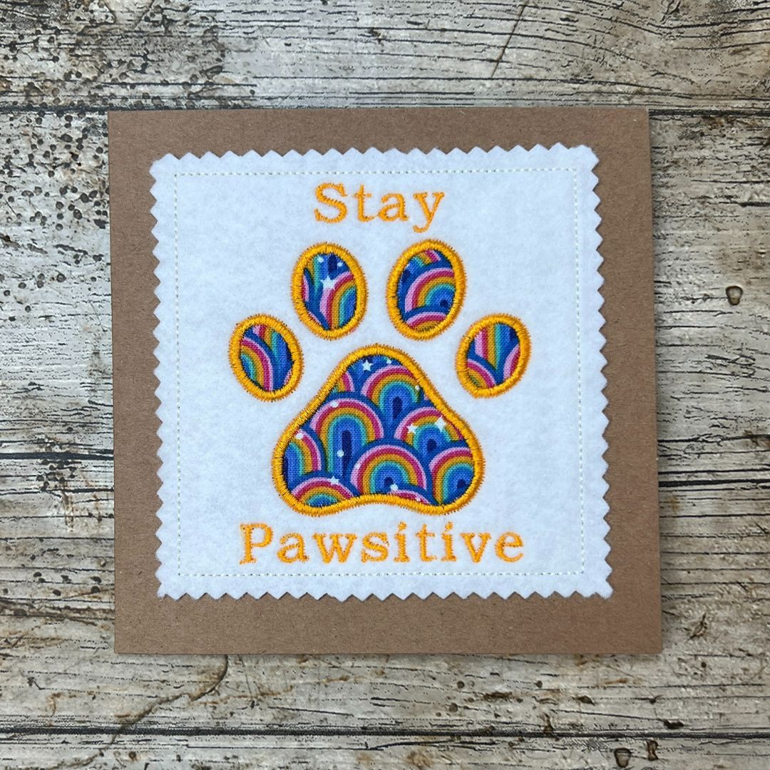 Card with rainbow paw print and 'Stay Pawsitive' text on a wooden surface