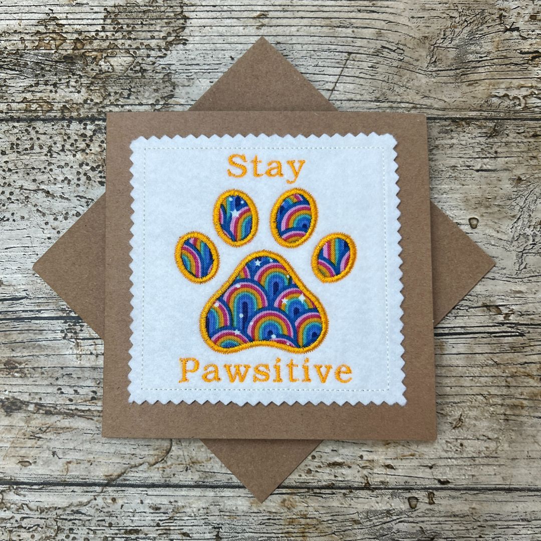 Card with rainbow paw print and 'Stay Pawsitive' text on a wooden surface