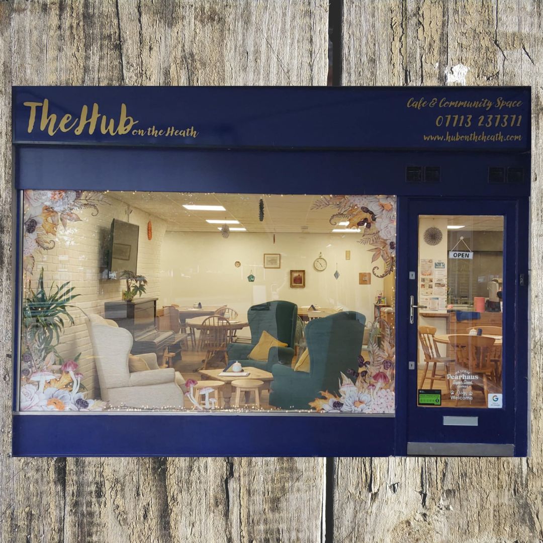 The Hub on the Heath cafe and community space storefront, featuring a dark blue facade, gold lettering, and a large display window showcasing cozy indoor seating and decor.