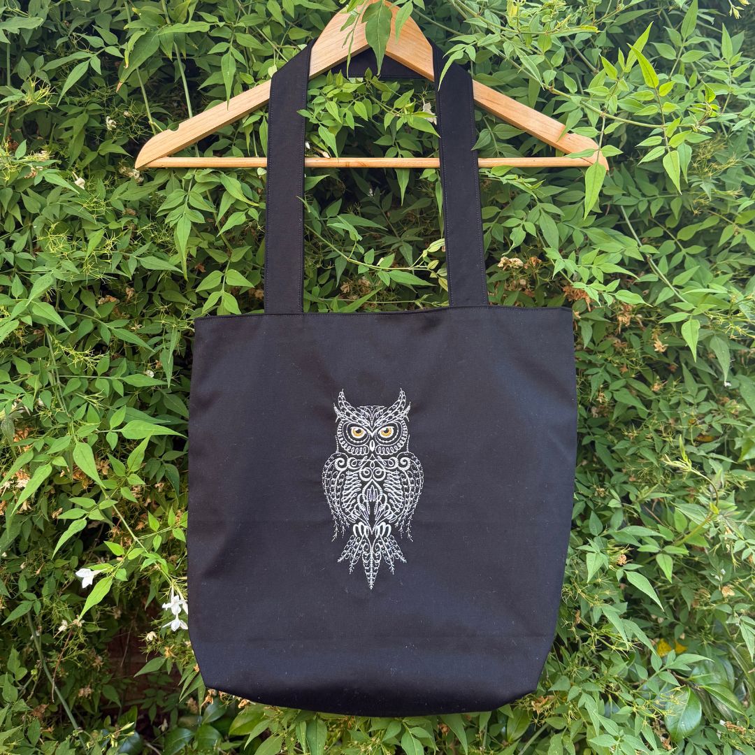 A black canvas tote bag with a white embroidered owl graphic hanging from a wooden hanger against a background of green foliage.