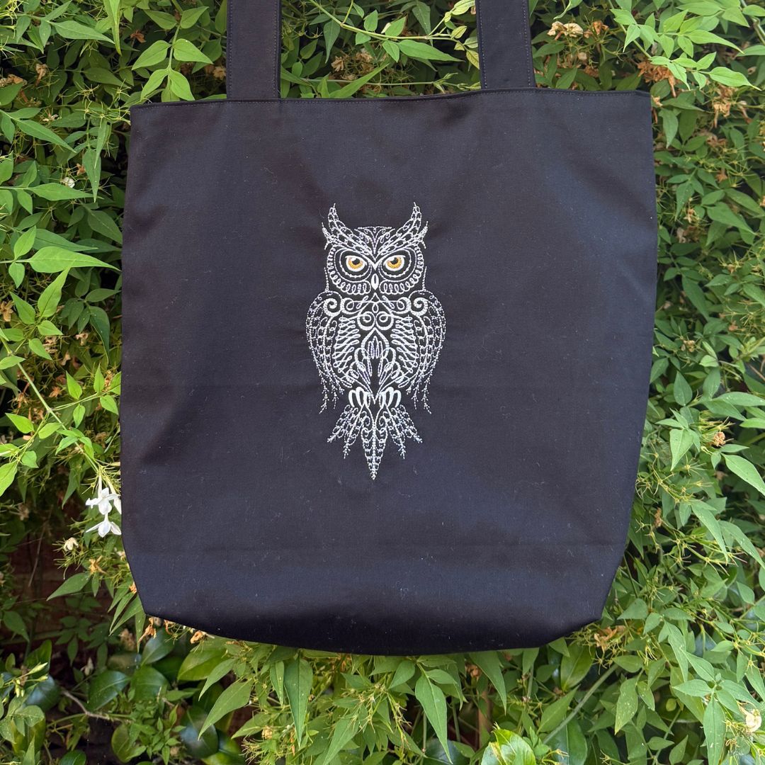 A black canvas tote bag with a white owl embroidered design hanging against a background of green foliage.