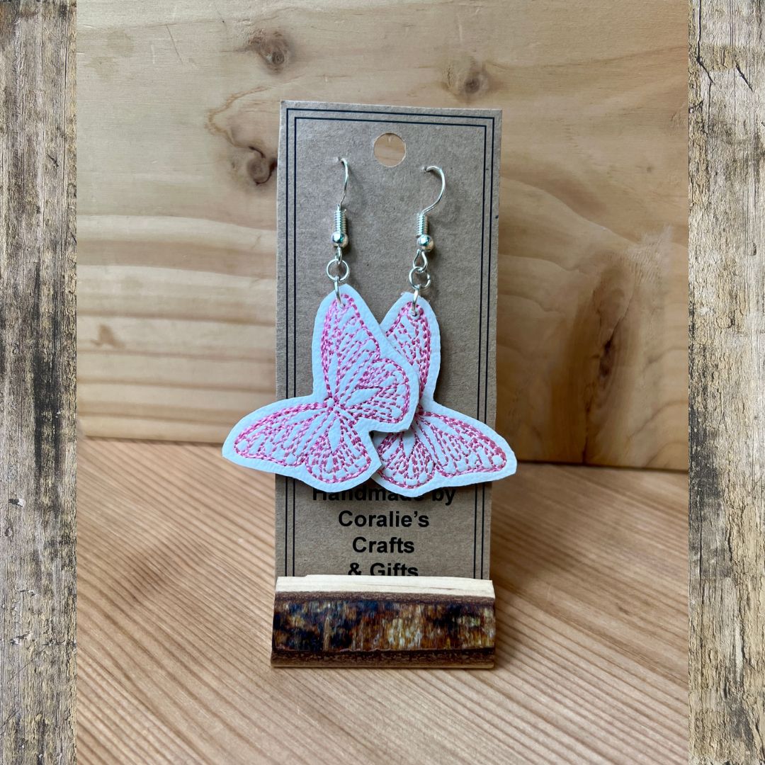 A pair of butterfly-shaped dangle earrings with pink embroidered details on a light blue background, hanging from silver hooks on a display card. The card is set against a rustic wooden backdrop.
