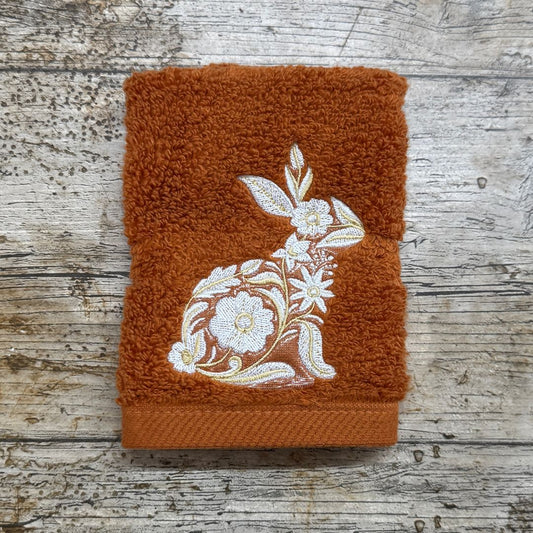 A folded terracotta orange face cloth with a white embroidered floral rabbit design, displayed on a rustic weathered wood background.
