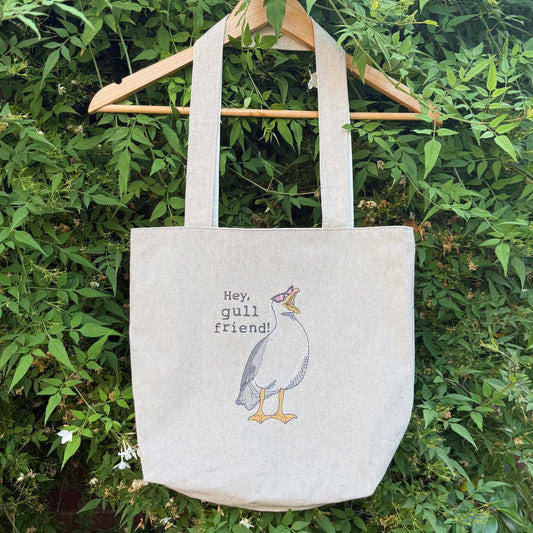 A natural canvas tote bag hanging from a wooden hanger against a background of green foliage. The bag features an embroidered illustration of a gull with the text 'Hey, gull friend!' on the front.