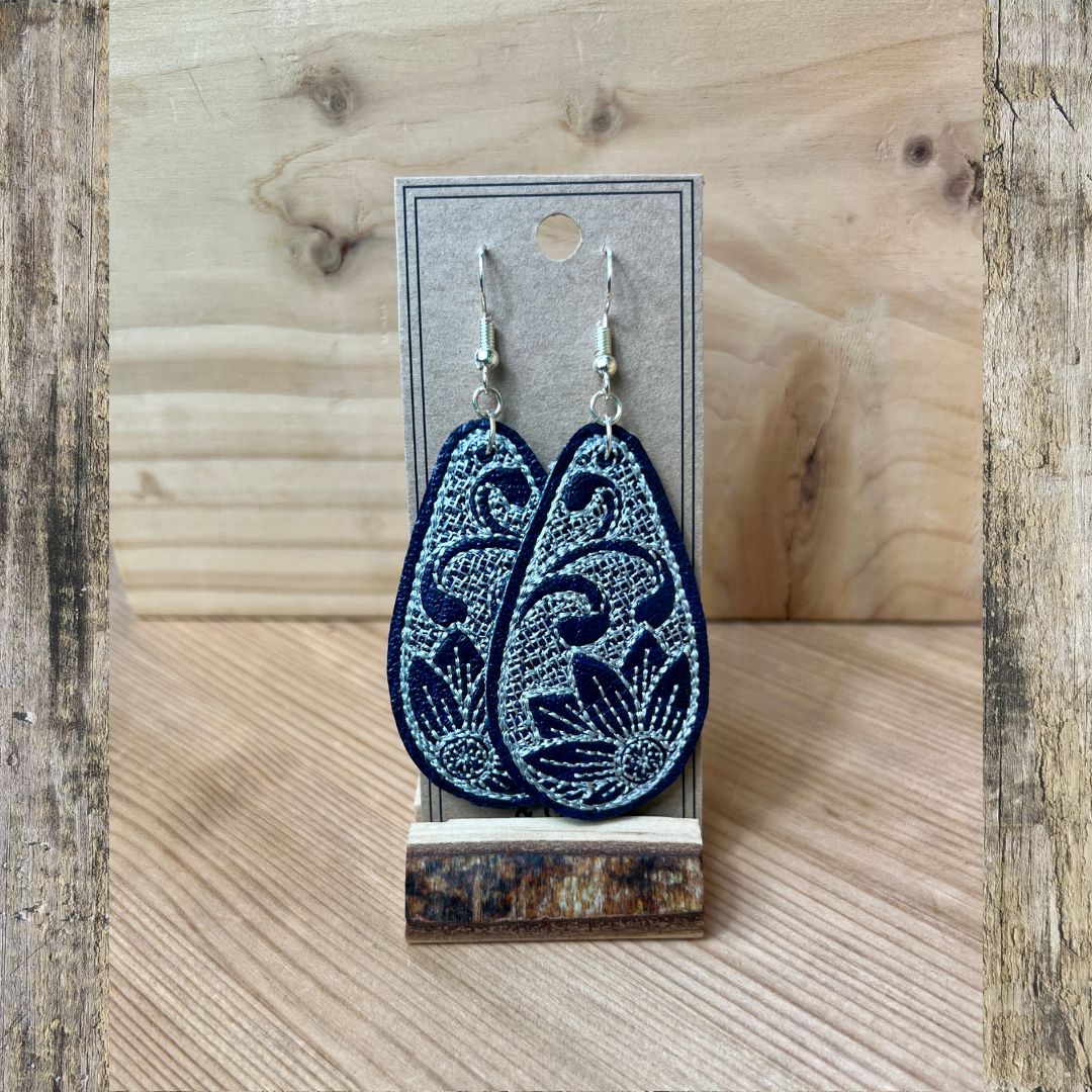 A pair of navy embroidered teardrop-shaped dangle earrings, with a white embroidered flower displayed on a gray card stand against a rustic wood background.