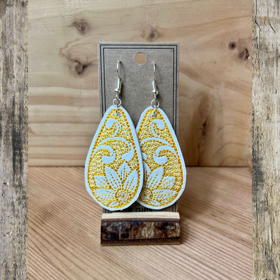 A pair of white embroidered teardrop-shaped dangle earrings, with a yellow embroidered flower displayed on a gray card stand against a rustic wood background.